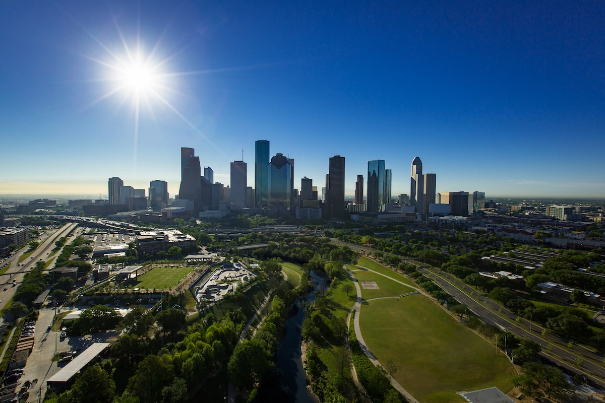 An aerial view of Houston's skyline reveals a clear blue sky with the bright sun shining above. In the foreground, lush greenery and a winding river create a picturesque scene, while towering skyscrapers stand proudly in the distance.