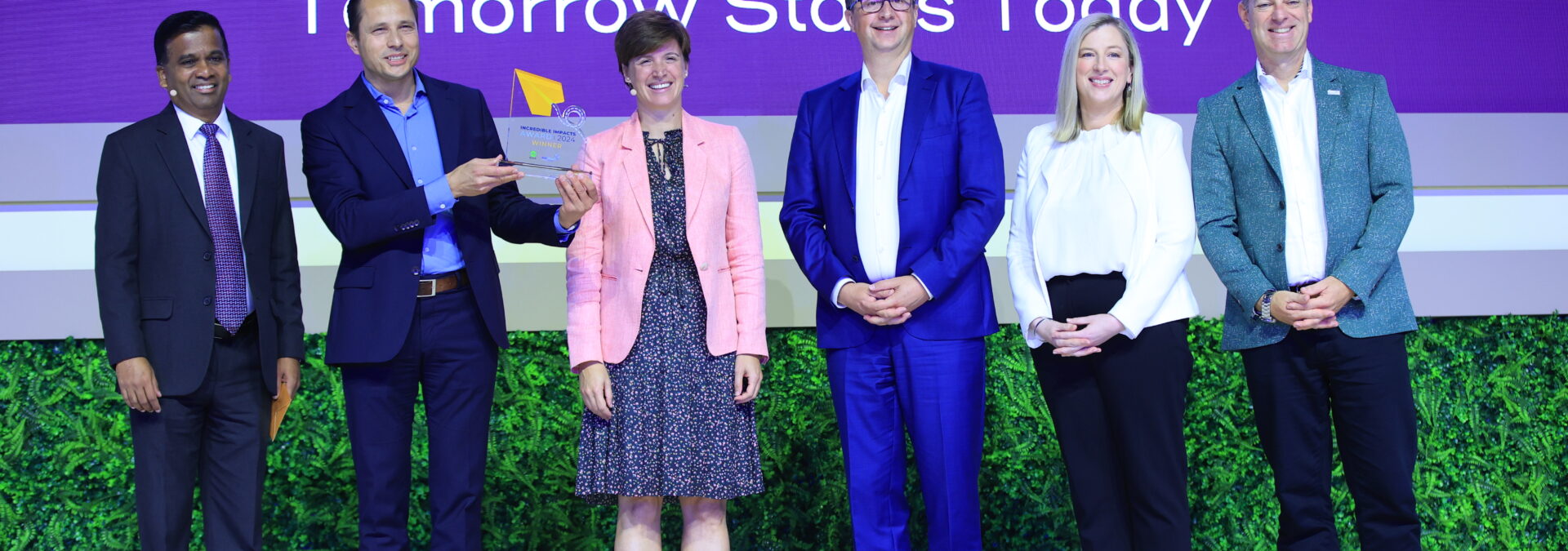 Six people stand on stage, three men and three women, posing for a group photo with one man holding a plaque. The background is purple, displaying the text 